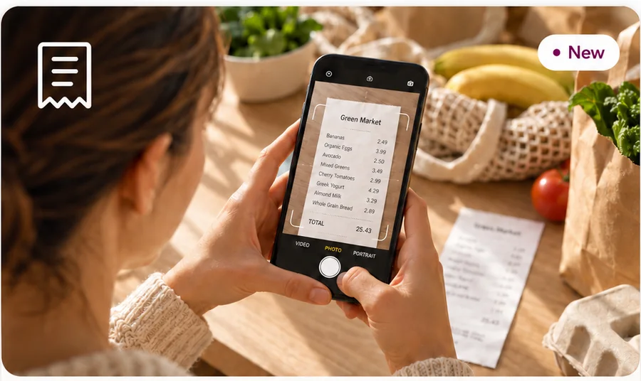 Someone photographing a grocery receipt with their phone, shopping bags and produce on the table beside them.